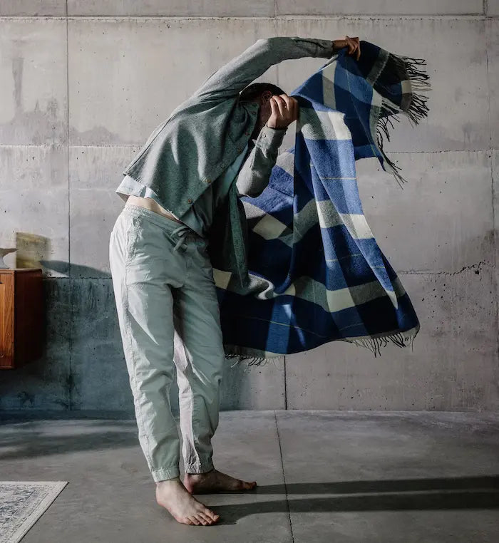 Man in neutral clothing, mid motion, swinging a blue, white and black tartan lambswool throw through the air