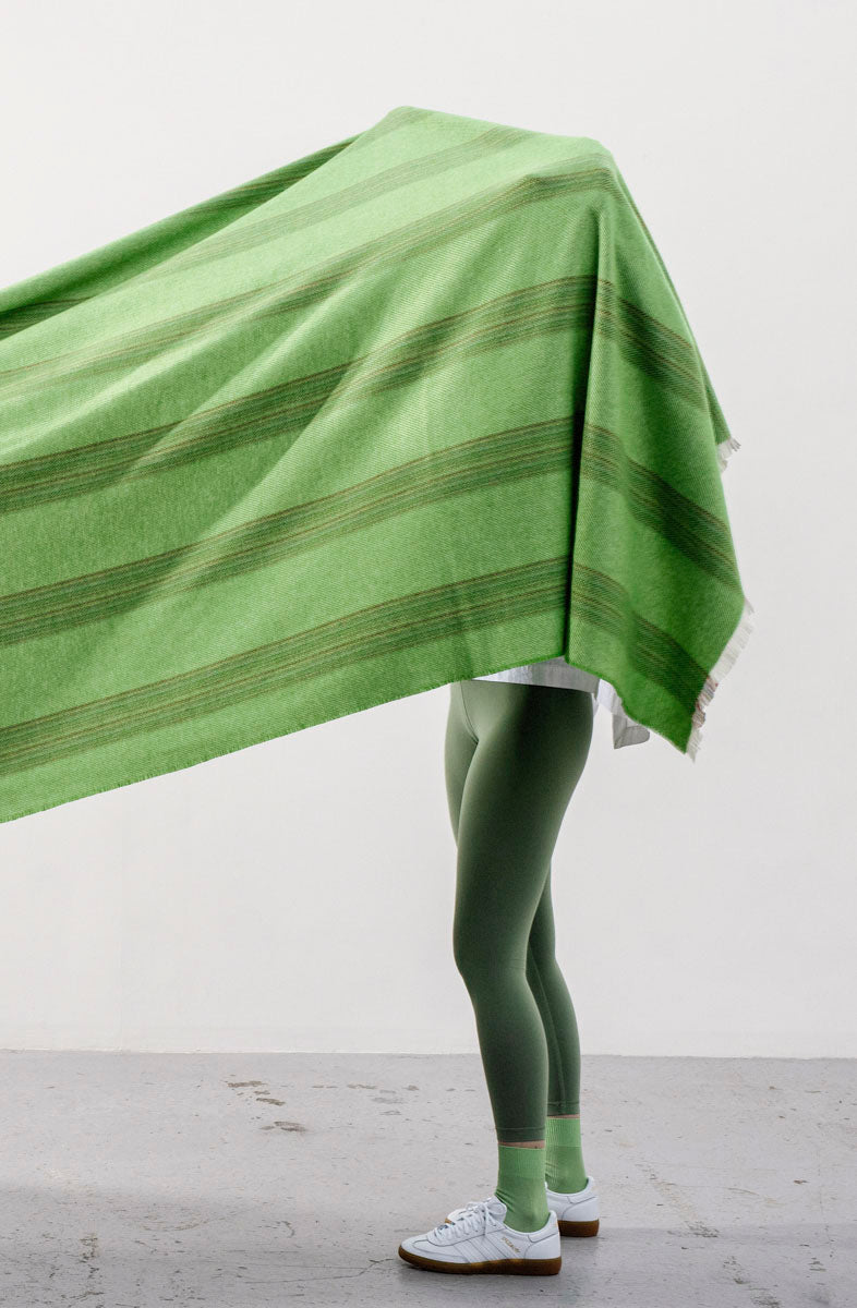 Person in green clothing holding up a light green striped blanket that covers their upper body 