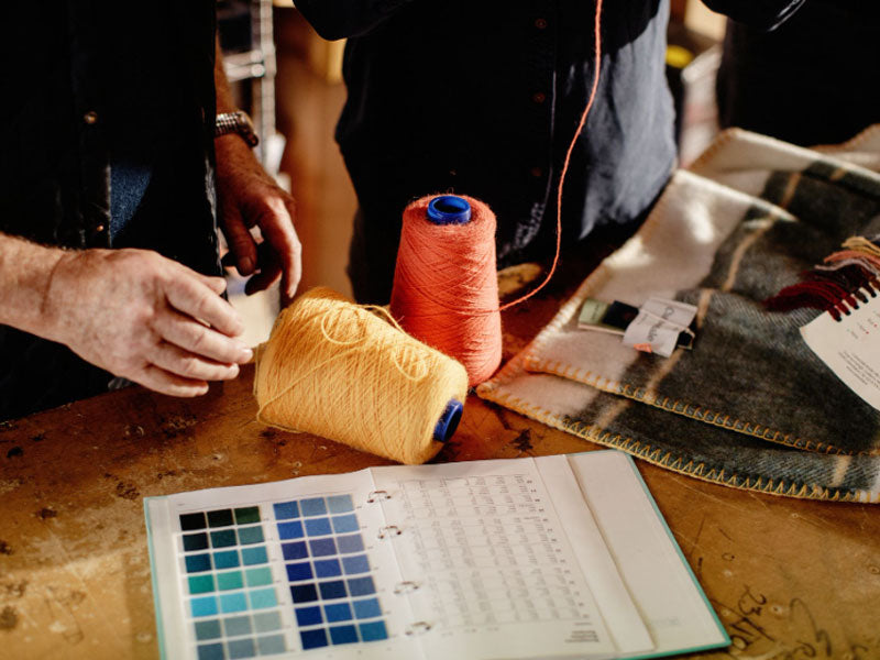 Figures standing over a worktable with colour swatches and spools of threads repairing blankets