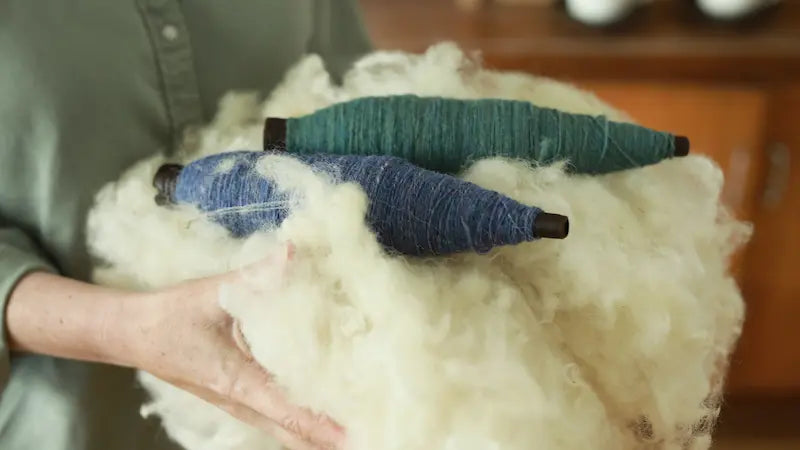Hands holding a bundle of sheared wool with spools of blue and green wool on top