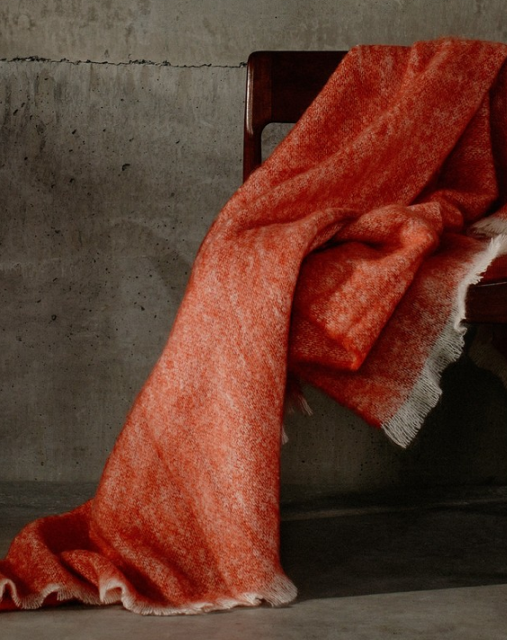 Soft dark orange woollen blanket draped over a dark wood chair in against a concrete wall
