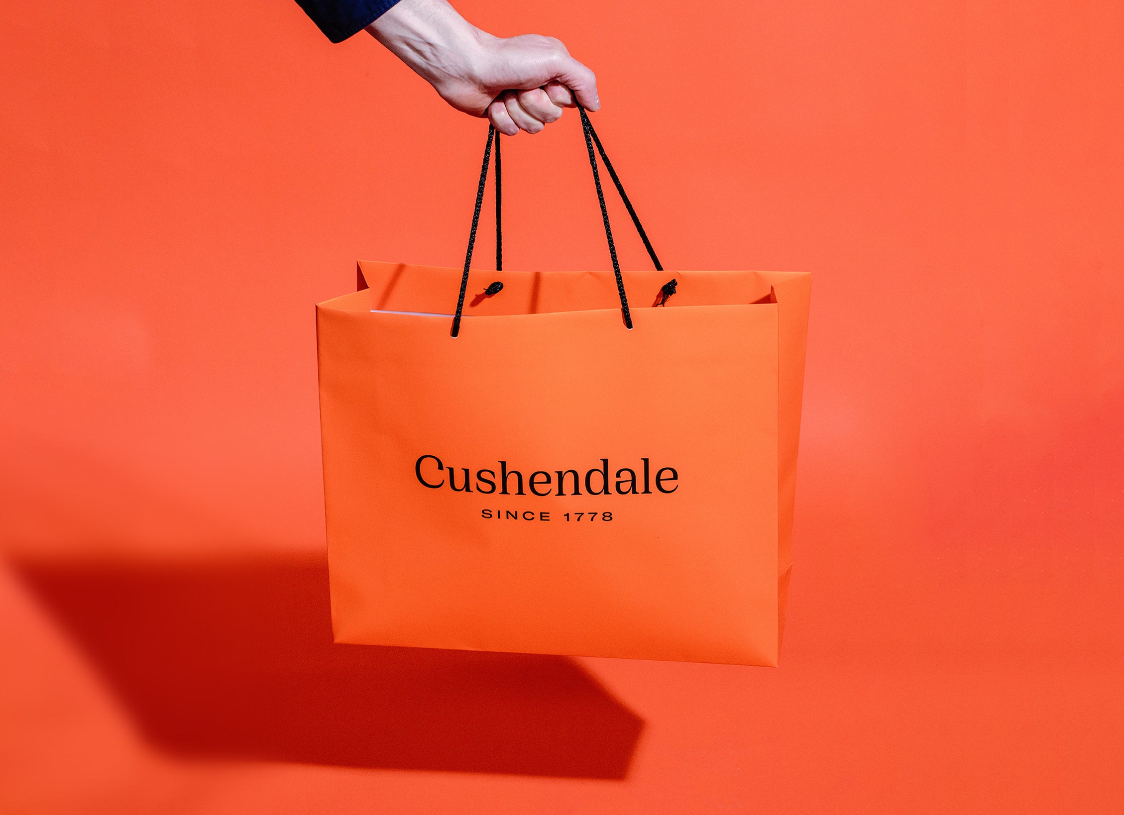 Hand holding a bright orange paper Cushendale shopping bag, against a bright orange background